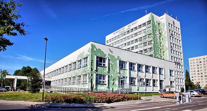 Socmodernistyczny dwubryłowy budynek w jasnych barwach z zielonym, roslinnym muralem zdobiącym elewację.