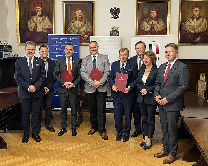 Ośmioosobowa grupa osób pozuje do zdjęcia. Trzech mężczyzn w środku trzyma bordowe teczki. W tle obrazy z pocztem rektorów.