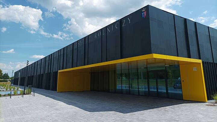 Nowoczesny podłuzny, dwukondygnacyjny budynek o grafitowej elewacji i zaznaczonym żółtym obrysem wejściem.